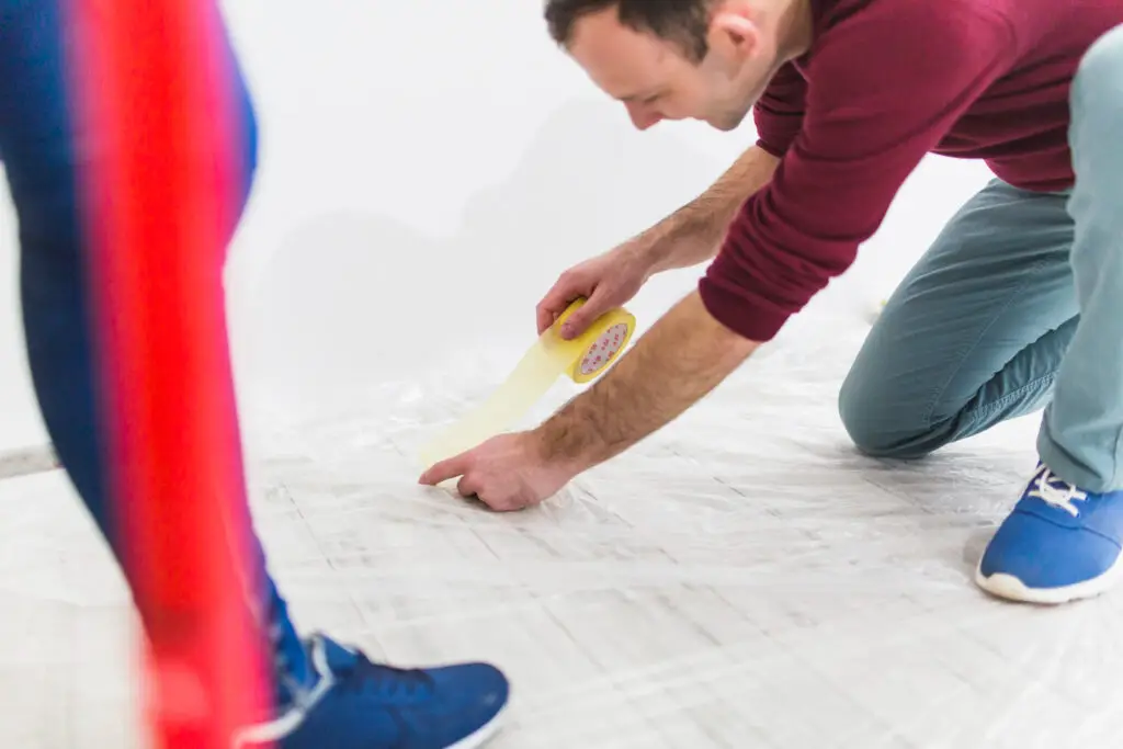 A person kneels on the floor applying tape, while another person stands nearby, both wearing casual clothing.