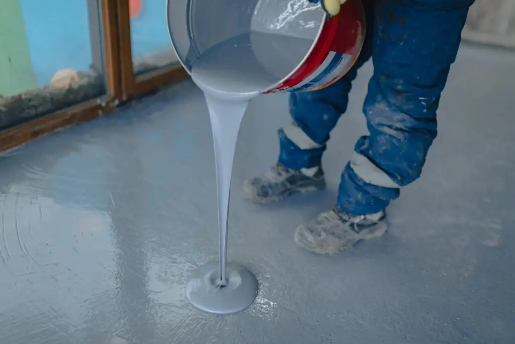 A person pours gray liquid cement mix from a red can onto a floor, wearing protective gear and work boots.