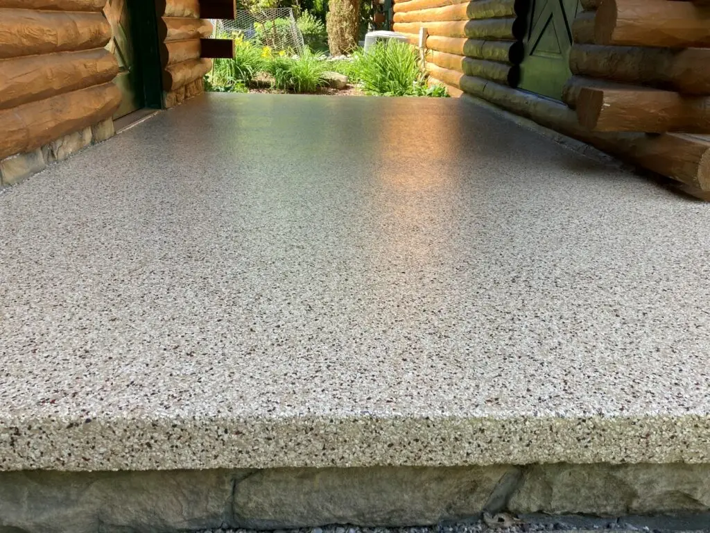 Textured concrete pathway between two log cabin walls, leading to a garden with vibrant greenery and sunlight filtering through the trees.
