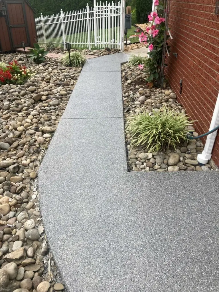 A well-maintained concrete path bordered by decorative stones and plants, leading to a white fence with a lush garden in the background.