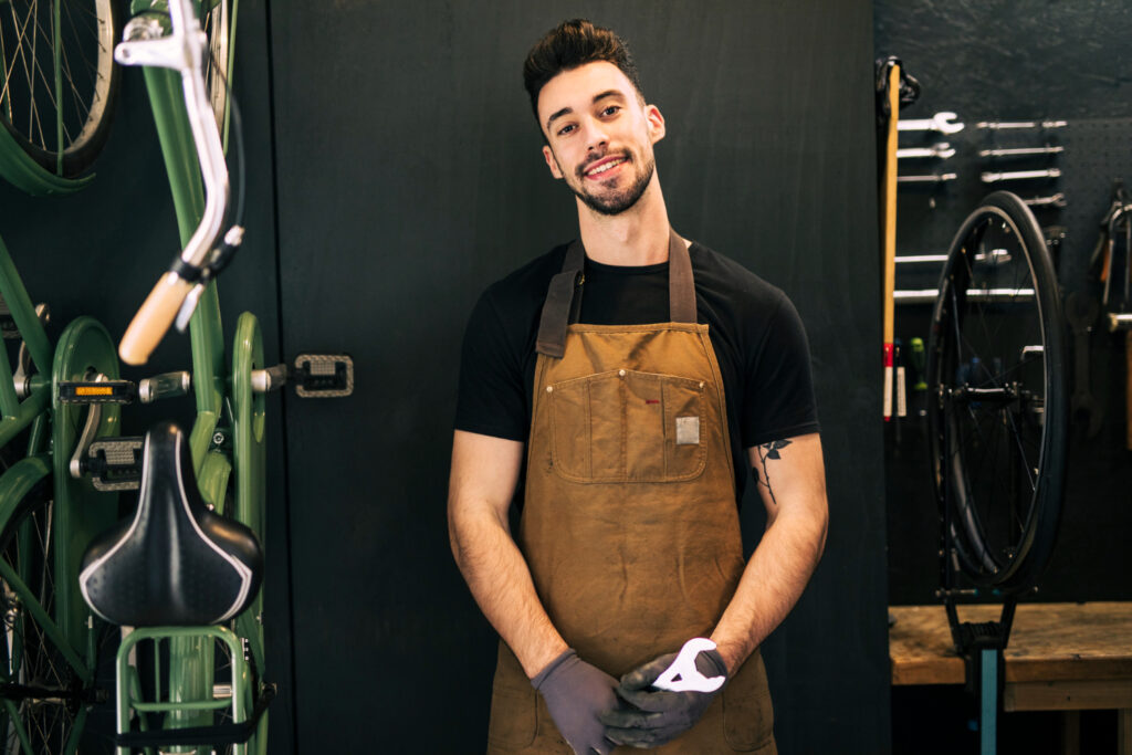 A person in a workshop, wearing an apron and gloves, stands beside tools and bicycles, appearing ready for mechanical work.