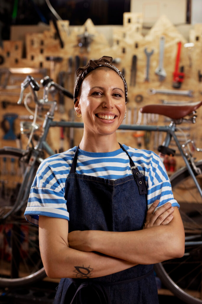 A smiling person in a striped shirt and apron stands in a workshop, surrounded by a bicycle and tools on a wall.