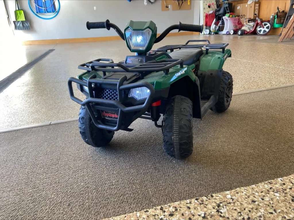 A child's green toy quad bike sits on a carpeted floor inside a garage, surrounded by various children's toys.