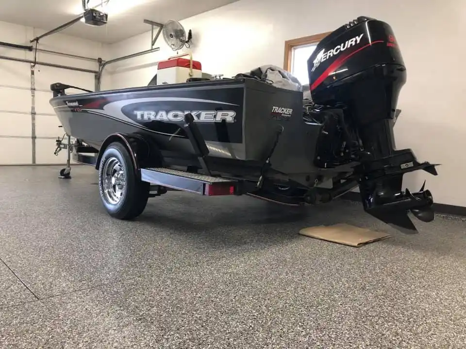 A black and red Tracker boat with Mercury engine is parked in a garage with a fan and overhead door.