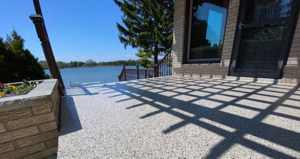 A sunlit patio overlooks a calm lake, surrounded by trees under a clear blue sky. The area features brickwork and metal railings.