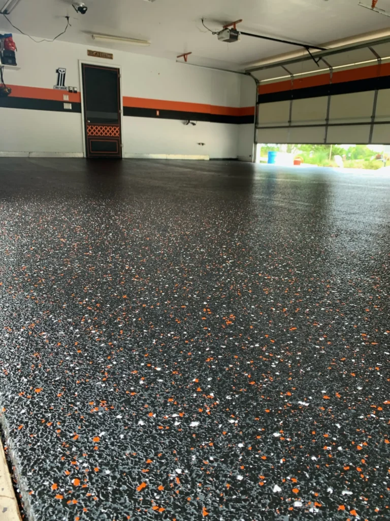 A garage with a speckled epoxy floor, white and orange wall stripe, closed door, and partially open garage door revealing greenery outside.