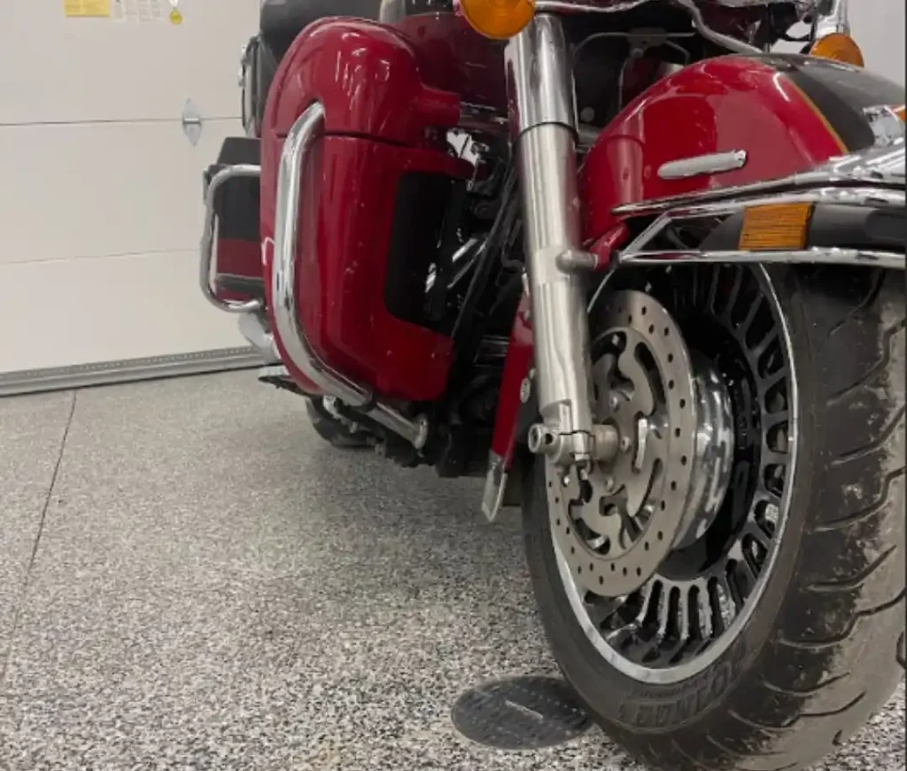 A red motorcycle with chrome details is parked indoors on a gray speckled floor, featuring visible front forks and tires.