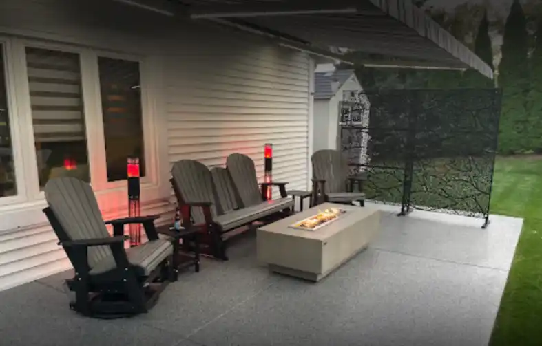 A cozy outdoor patio features Adirondack chairs, fire pit table, retractable awning, and privacy screen, surrounded by lush greenery, creating a relaxing atmosphere.