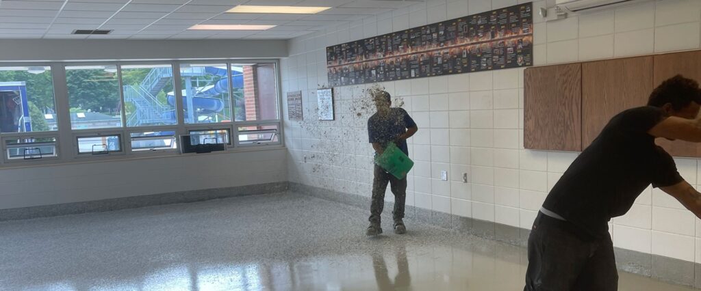 Two people in a room, one splashing water from a bucket. Windows show an outdoor play structure. The walls have decorative panels.