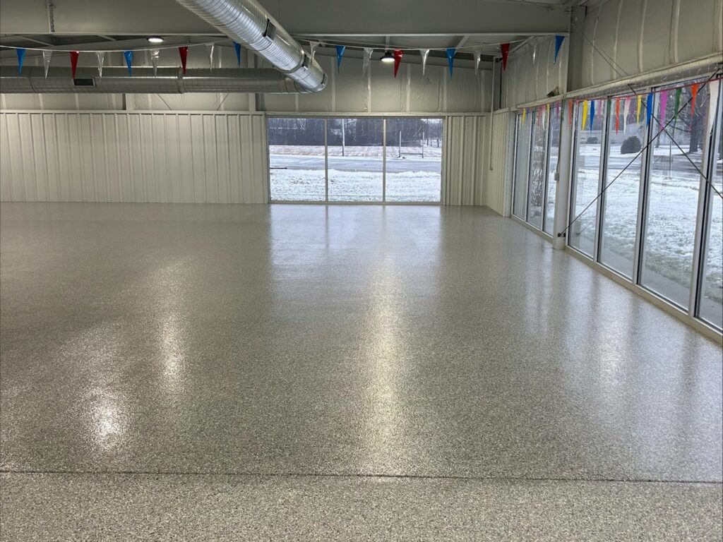 Empty industrial space with polished concrete floor, colorful pennant banners, large windows, and snowy view outside. Ducting visible on ceiling.