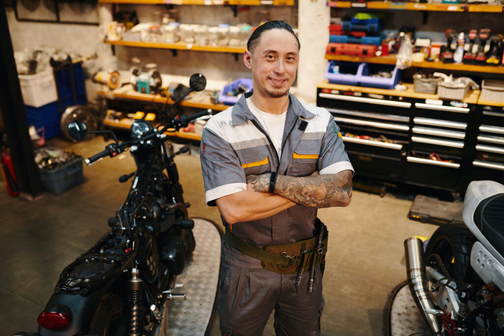 A person in a uniform stands confidently in a motorcycle workshop, surrounded by tools and vehicles, with arms crossed and smiling.