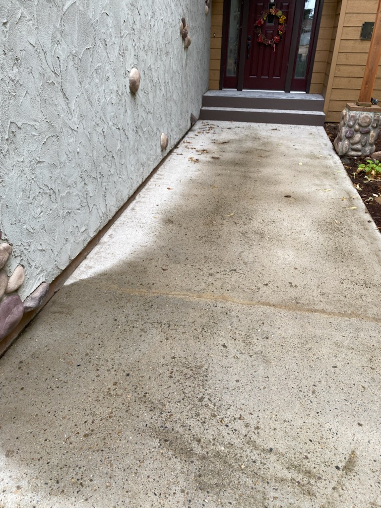 Concrete pathway leading to a front door with a fall-themed wreath, bordered by textured and wooden walls, surrounded by stones.