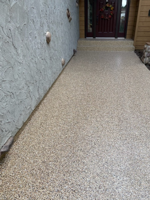 A textured stone pathway leads to a wooden door adorned with a floral wreath, flanked by a stucco wall and wooden siding.