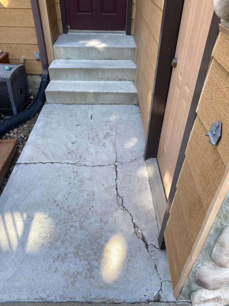Concrete steps with visible crack on the walkway lead to a door, surrounded by brown siding, near an air conditioning unit.