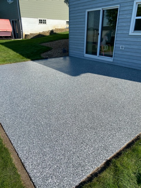 A newly installed grey concrete patio beside a house with sliding glass doors, surrounded by green grass and a neighboring building.