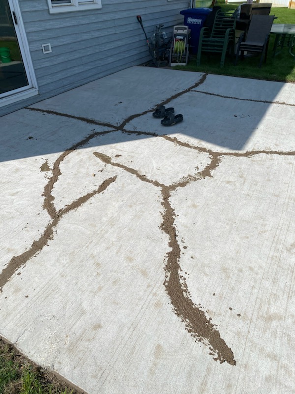 A patio with dirt-filled cracks, two shoes, stacked chairs, and a blue recycling bin near a house wall, under sunlight and shadows.